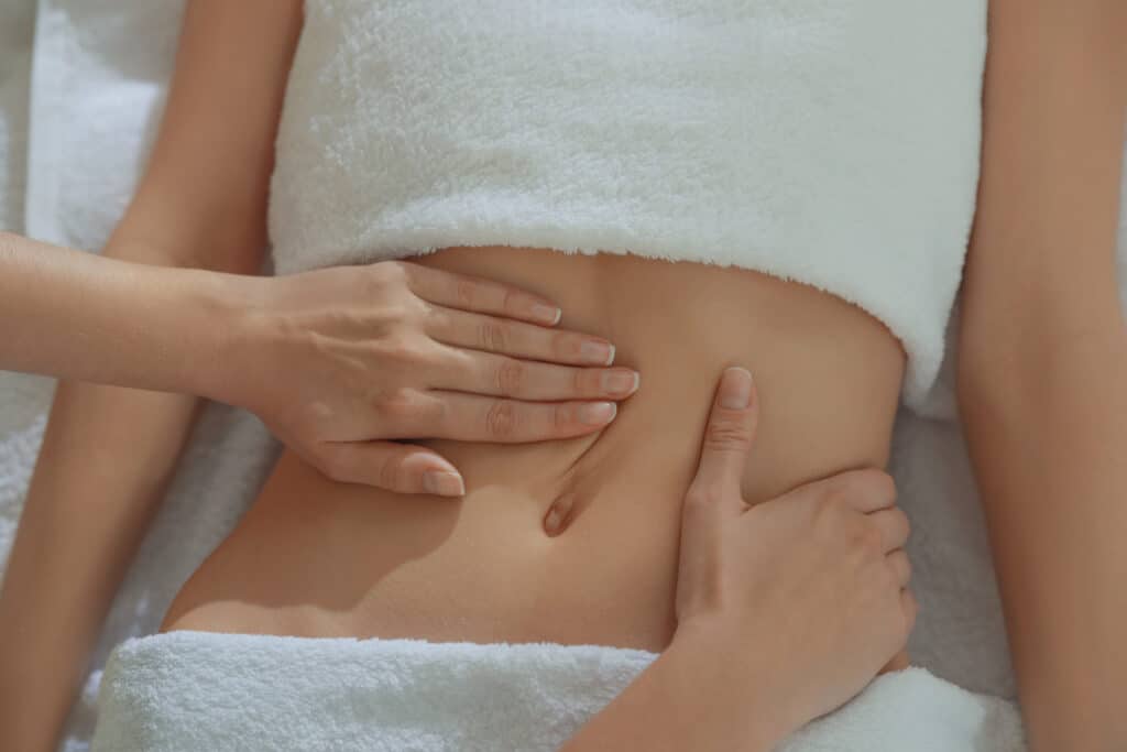 Mains expertes massant doucement l'abdomen d'une personne allongée, recouverte de serviettes blanches, favorisant la détente et le bien-être.